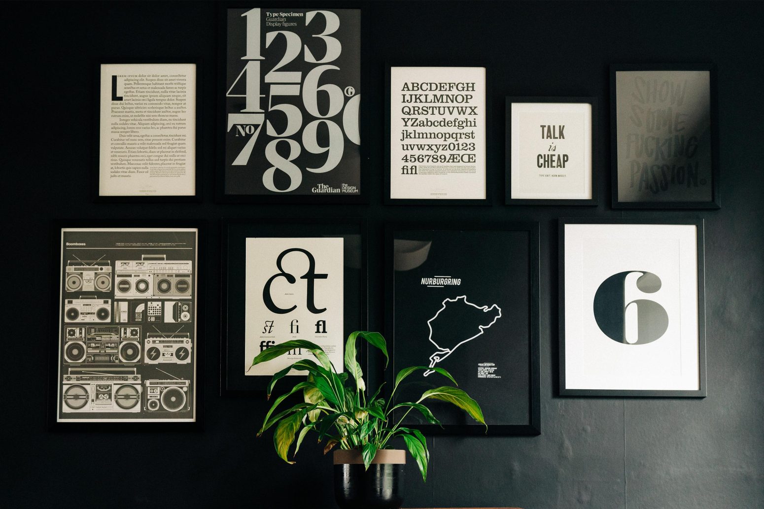 A dark wall adorned with various framed artworks featuring graphic design and symbols, alongside a potted plant on a shelf beneath the frames.