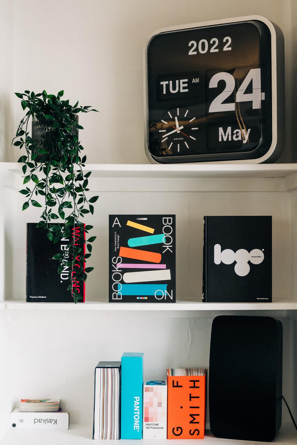 A modern indoor shelf displaying various design books, a large digital clock showing the date and time, and an indoor plant on the left. Emphasizing a minimalist aesthetic, the setup mirrors the brand design