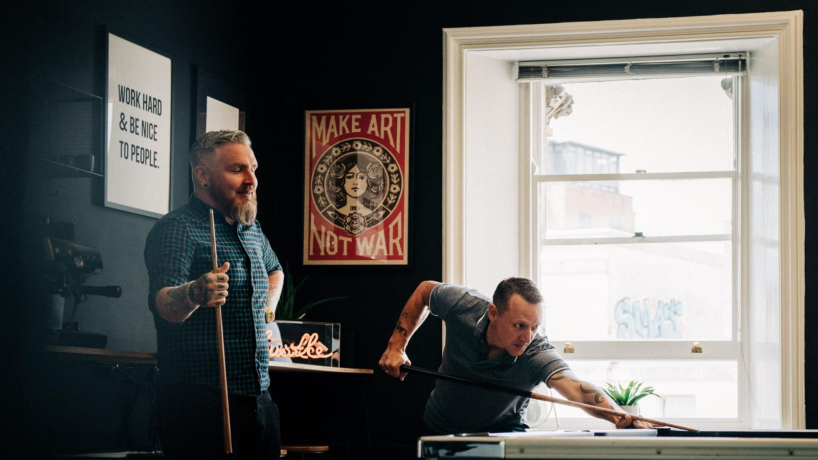 Two men playing pool in a room with dark walls adorned with motivational posters. One man, with a beard, watches while the other, wearing a dark t-shirt featuring a subtle 