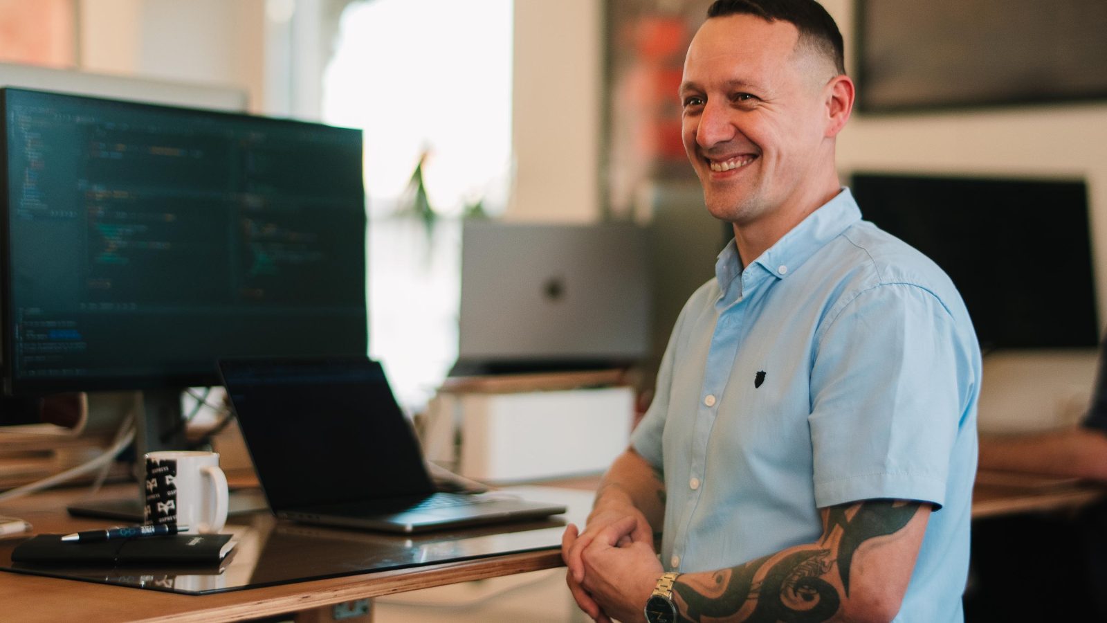 A smiling man with a sleeve tattoo stands at a desk with multiple computers in a modern Design Agency UK office.