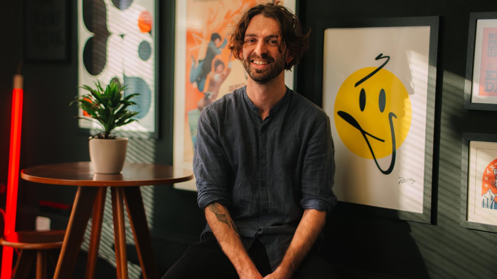 A smiling man with curly hair, dressed in a blue shirt, sits in front of brightly-colored graphic posters and a potted plant in a stylish room at a design agency in the UK.