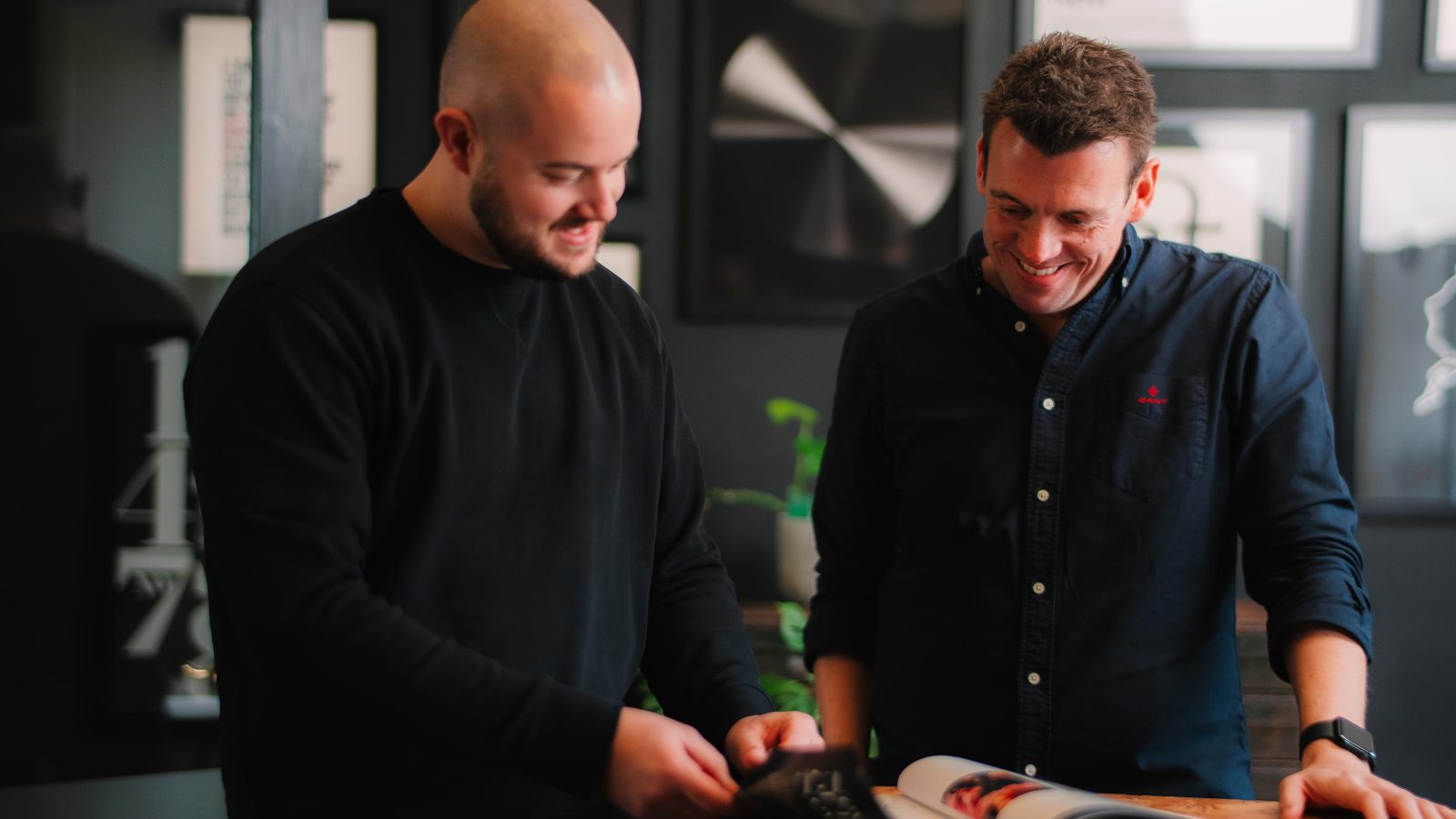 Two men, one bald and one with short hair, smiling and looking at a website design magazine together in a modern room with stylish decor.