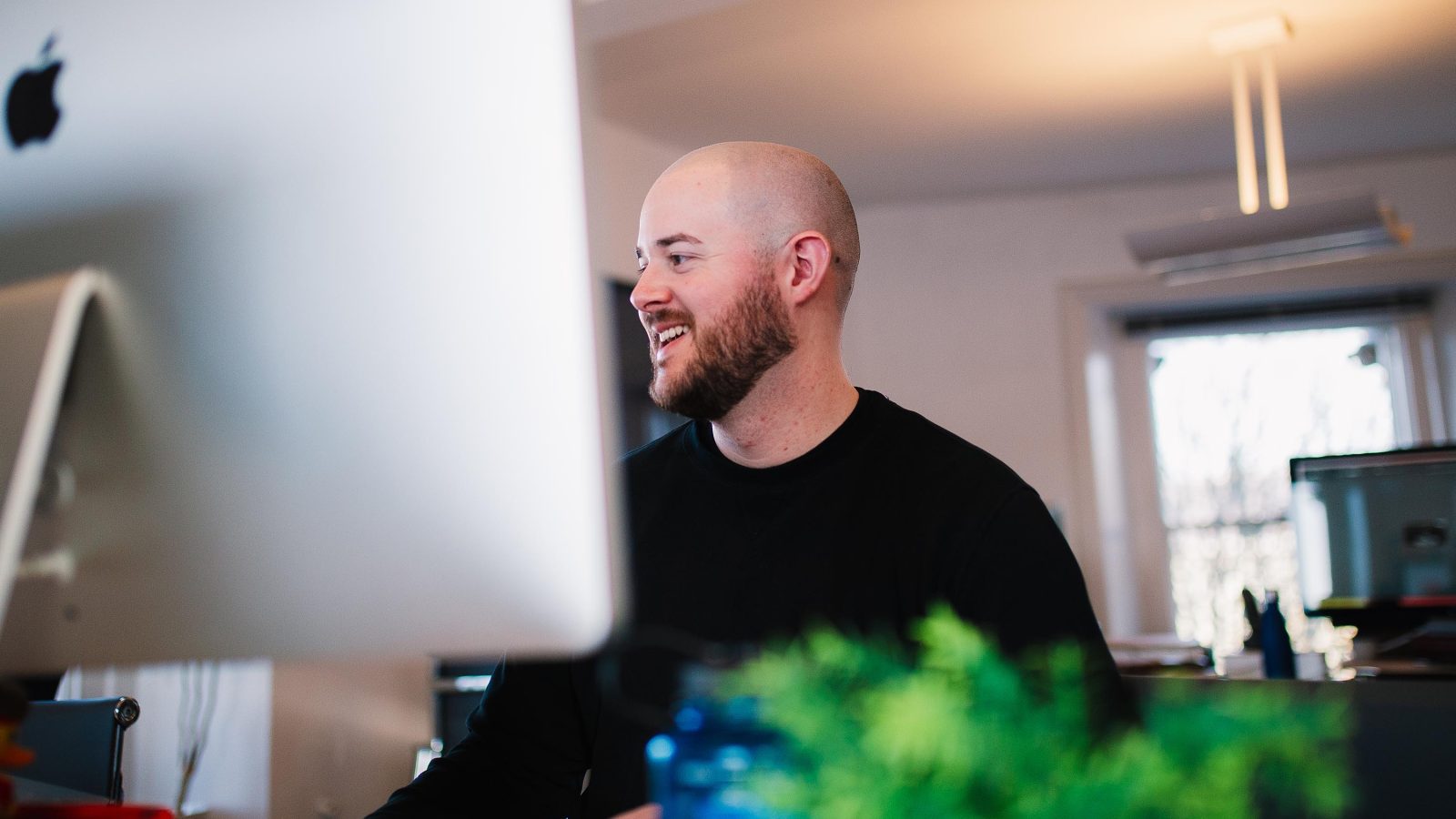 A bald man in a black shirt smiles while working on website design at a desk with an iMac and other office items in a well-lit room.