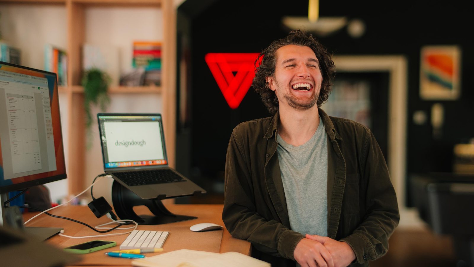 A joyful young man with curly hair laughing in an office setting, with a laptop displaying 