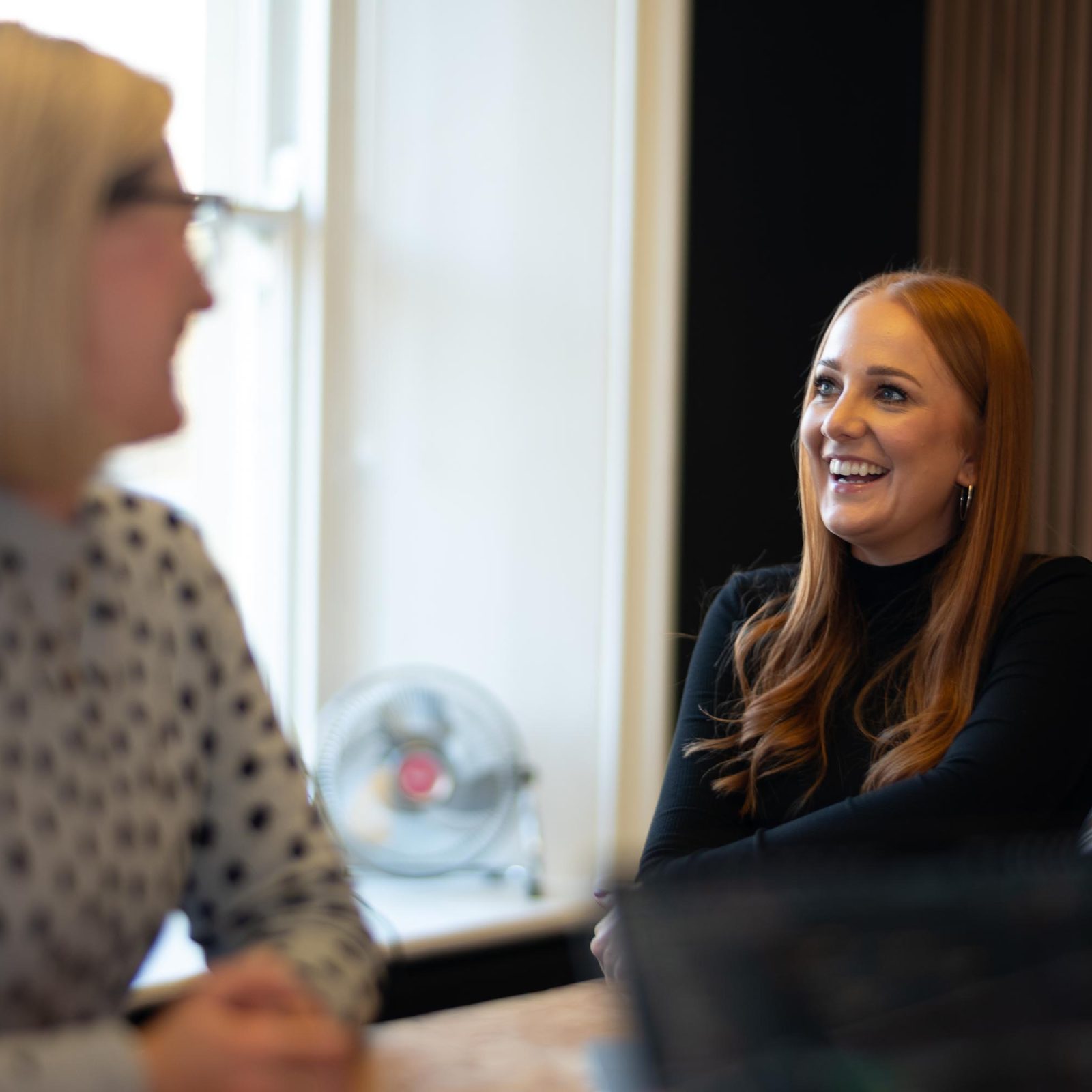 Two women engage in a joyful conversation at a work desk in a brand agency. One, with long red hair, smiles broadly at her blonde counterpart in a blurred foreground, reflecting a friendly, professional setting