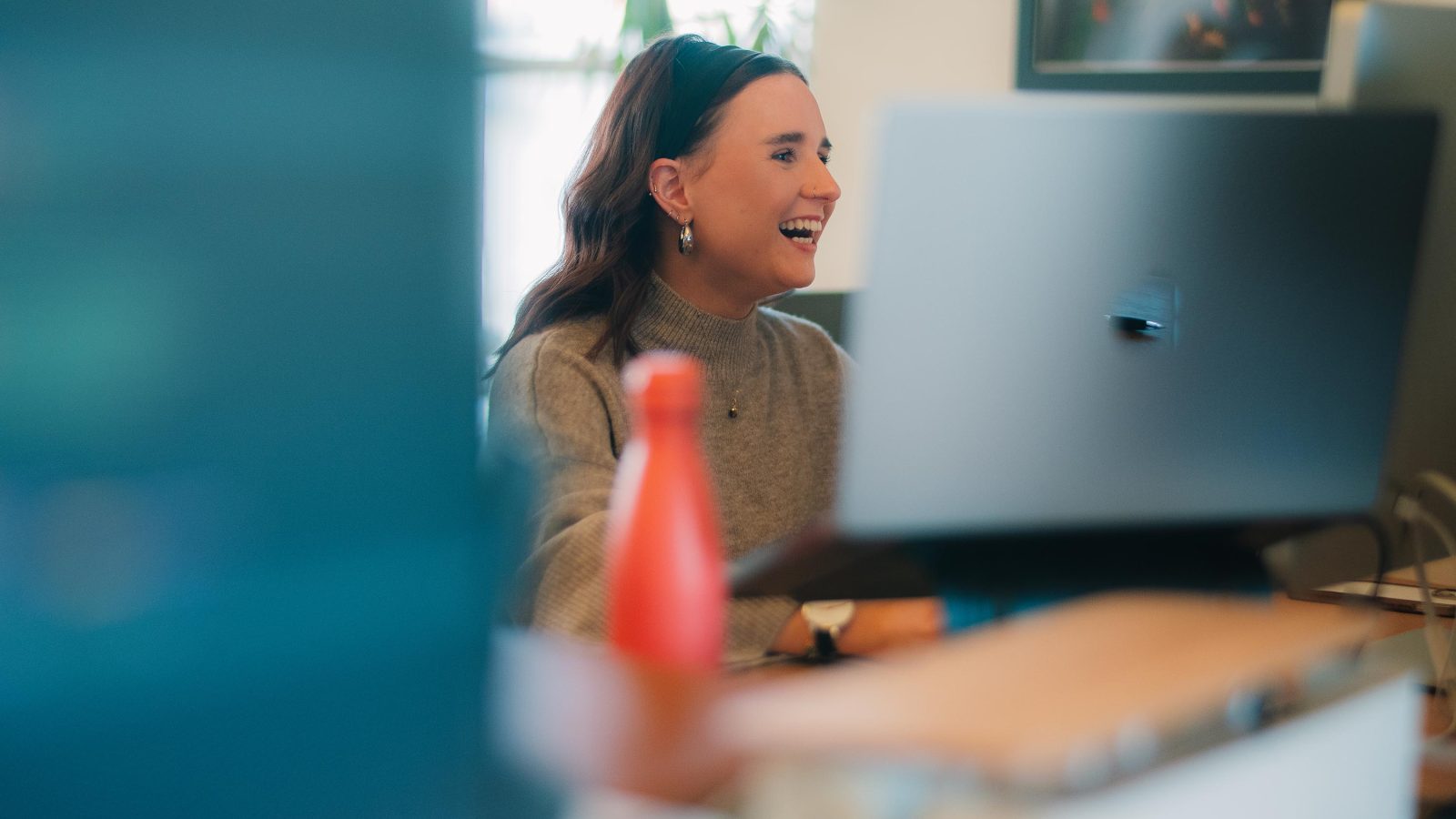 A smiling woman in a grey sweater works at a desktop computer in a brightly lit office of a Brand Agency UK, with a blurred out bottle in the foreground.