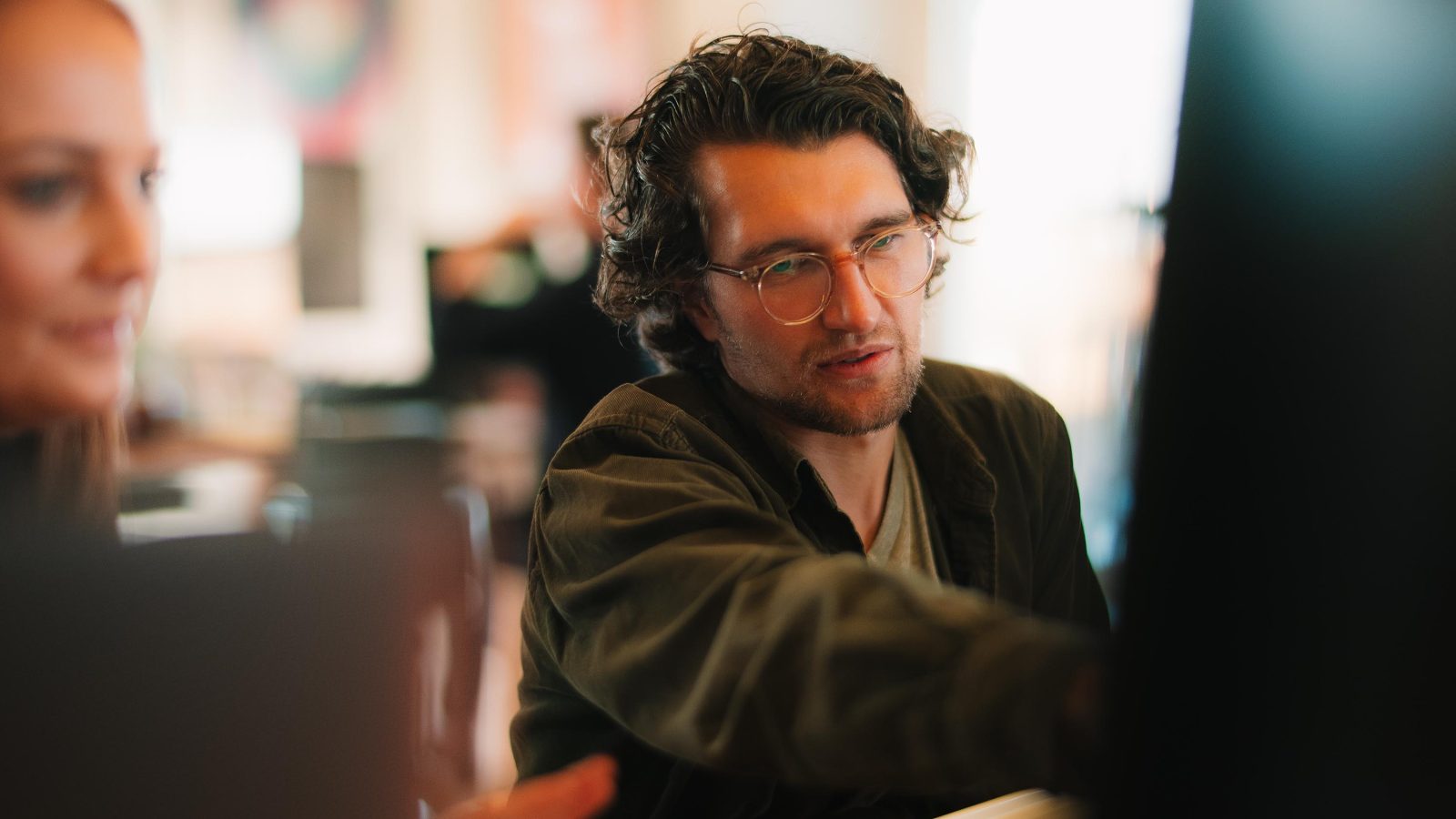 A young man with curly hair and glasses explaining something about brand design to a colleague, both focused on a computer screen in a brightly lit office.