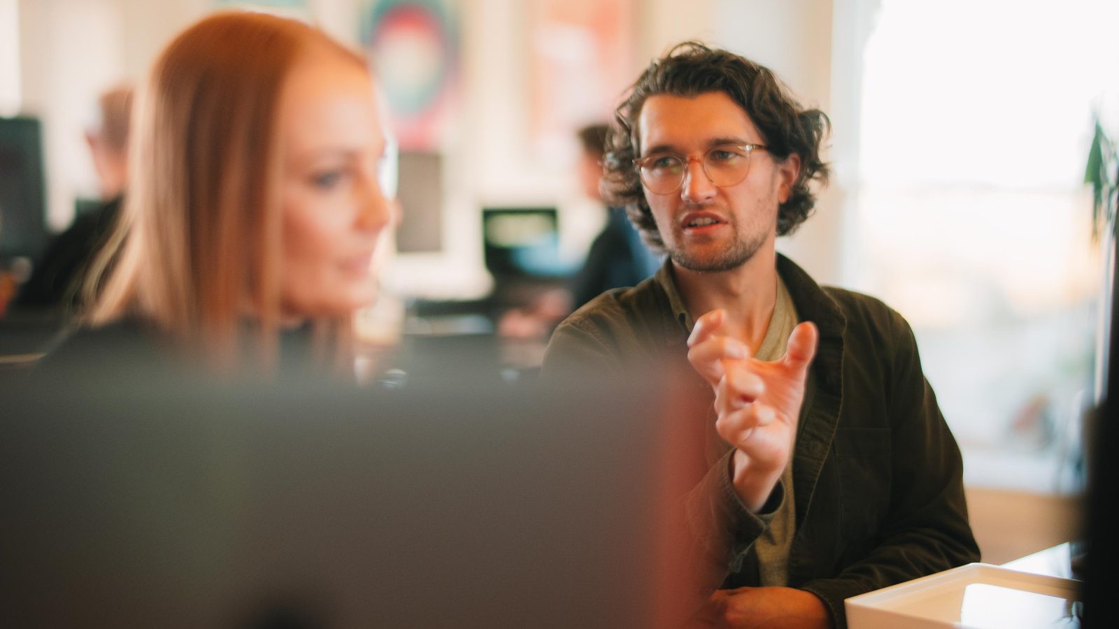 A man with curly hair gesturing while talking across the table to a focused redhead woman in an office setting, blurred computer screen displaying brand design in the foreground.
