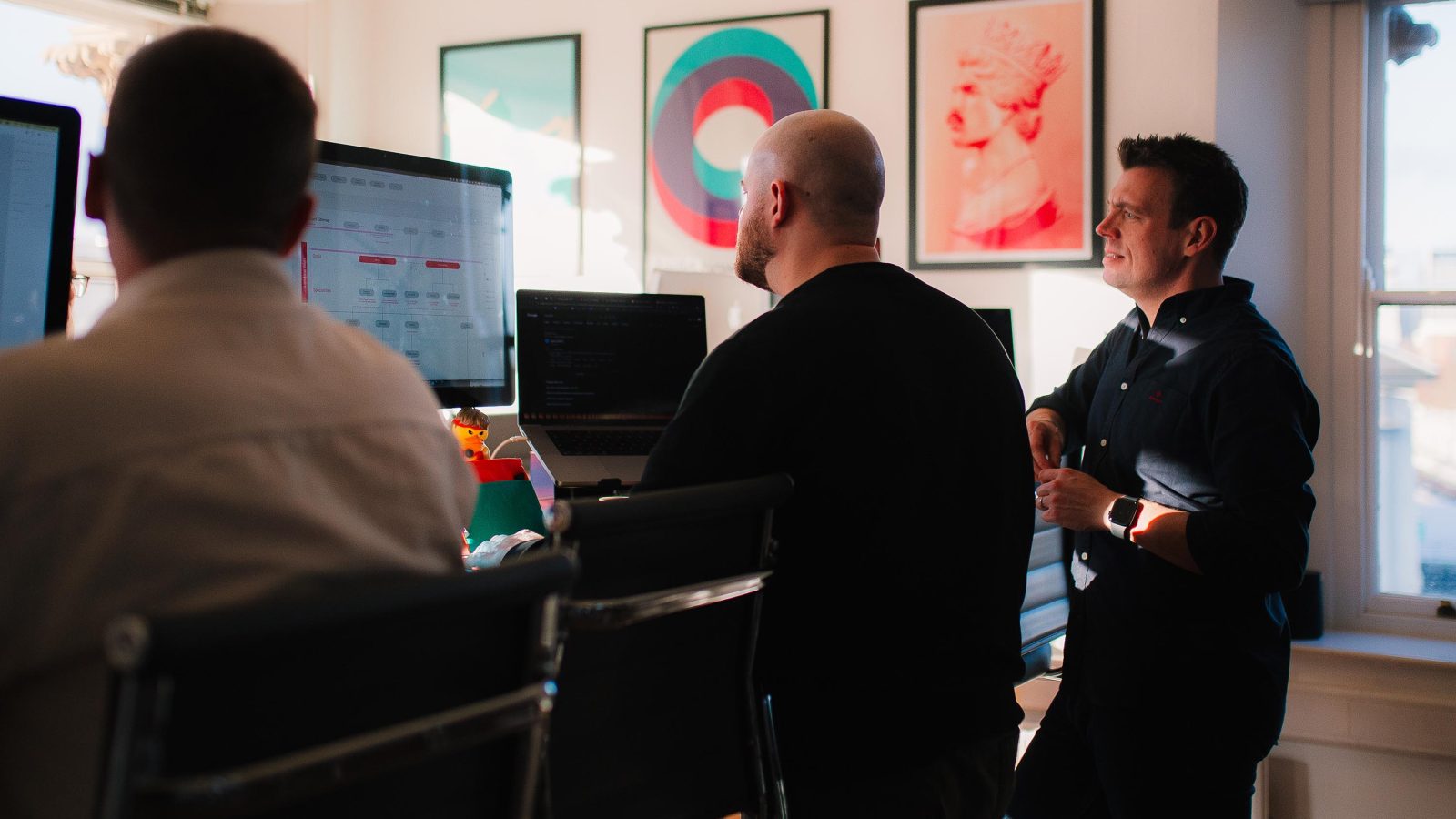 Three professionals in an office discussing brand design services in front of computer screens, with colorful artwork on the walls.