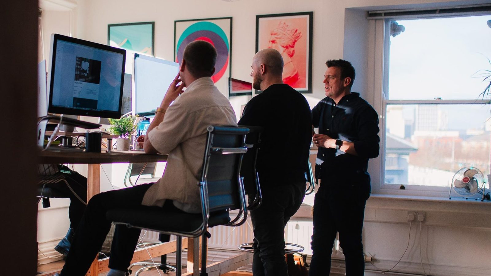 Three men in a sunny, artful office at a design agency; two are discussing careers while the third focuses on his computer work. The room is vibrant with wall art and large windows.