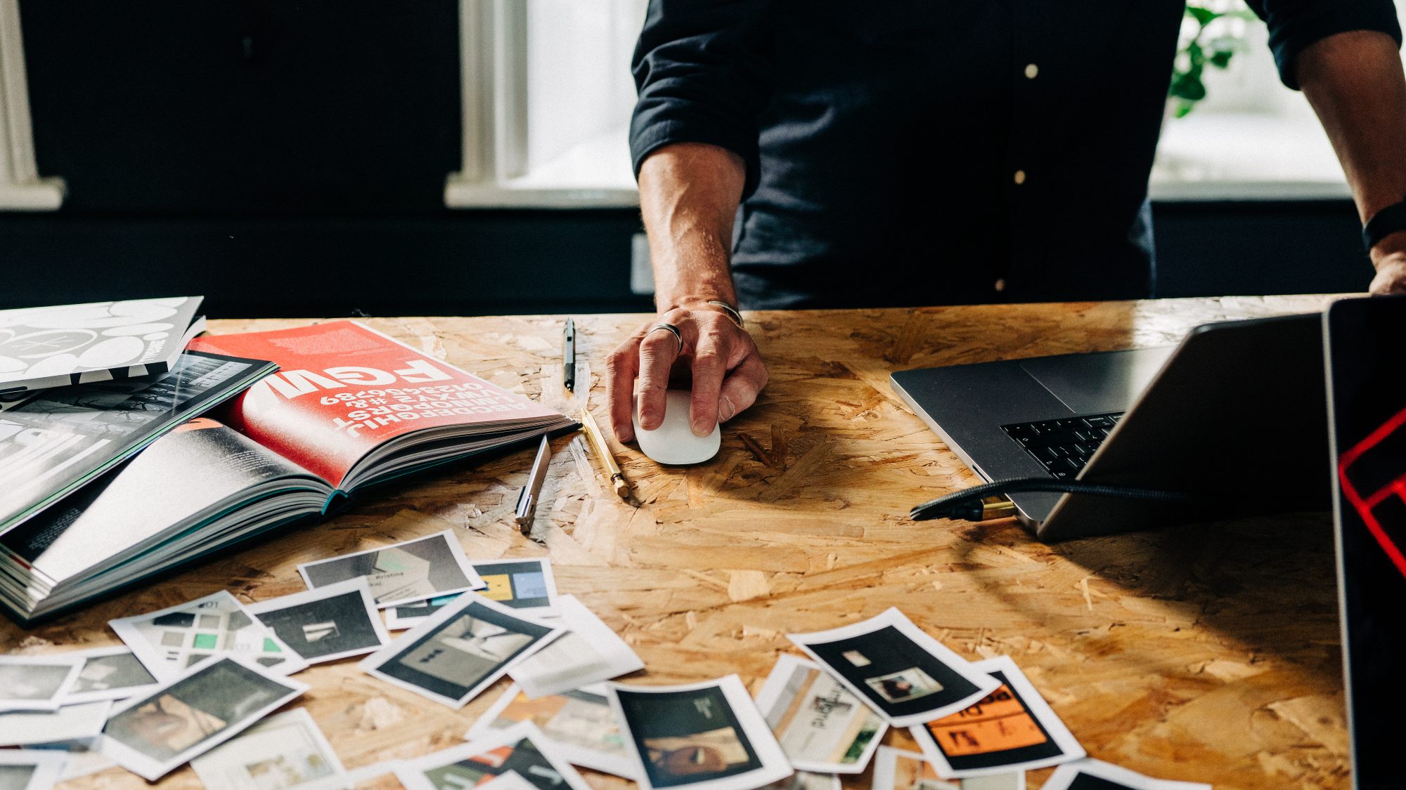 A person's hand using a computer mouse on a wooden desk, surrounded by scattered photographs and a laptop next to an open magazine, while working on website design.