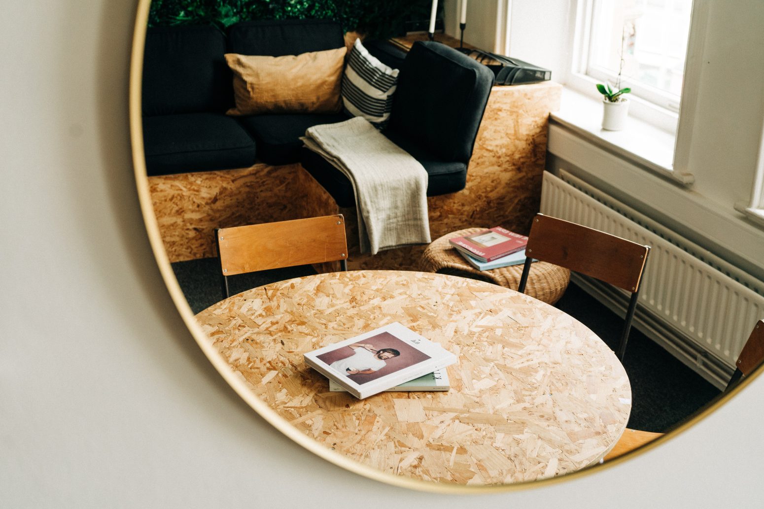 A cozy reading nook with a black sofa, wooden chairs, and a round chipboard table featuring a book on website design with a person on the cover, all reflected in a large circular mirror.