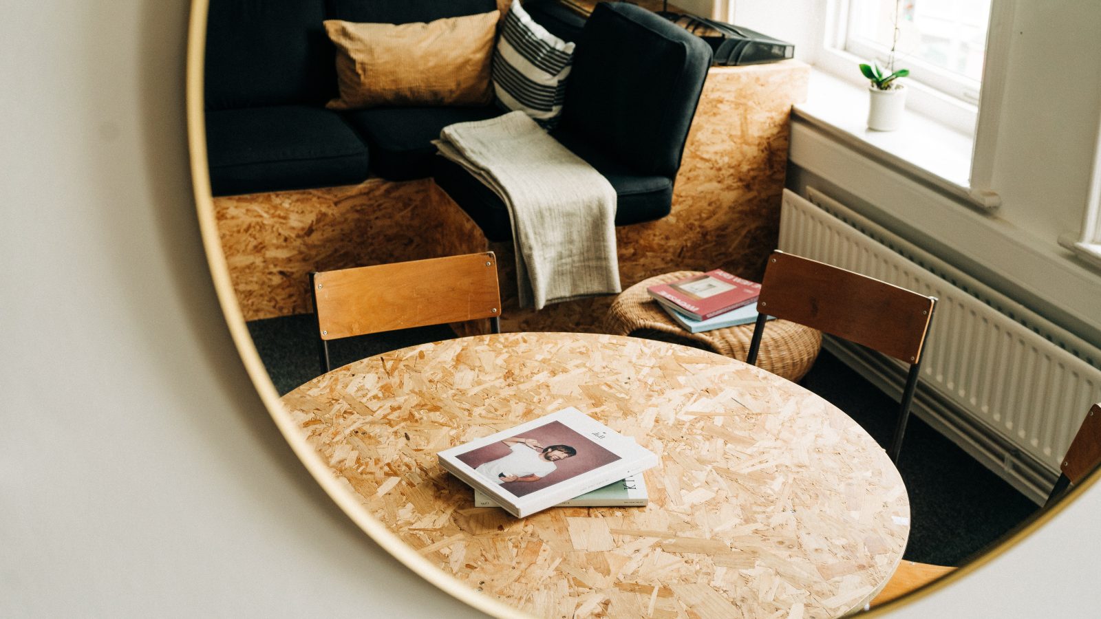 A cozy reading nook with a black sofa, wooden chairs, and a round chipboard table featuring a book on website design with a person on the cover, all reflected in a large circular mirror.