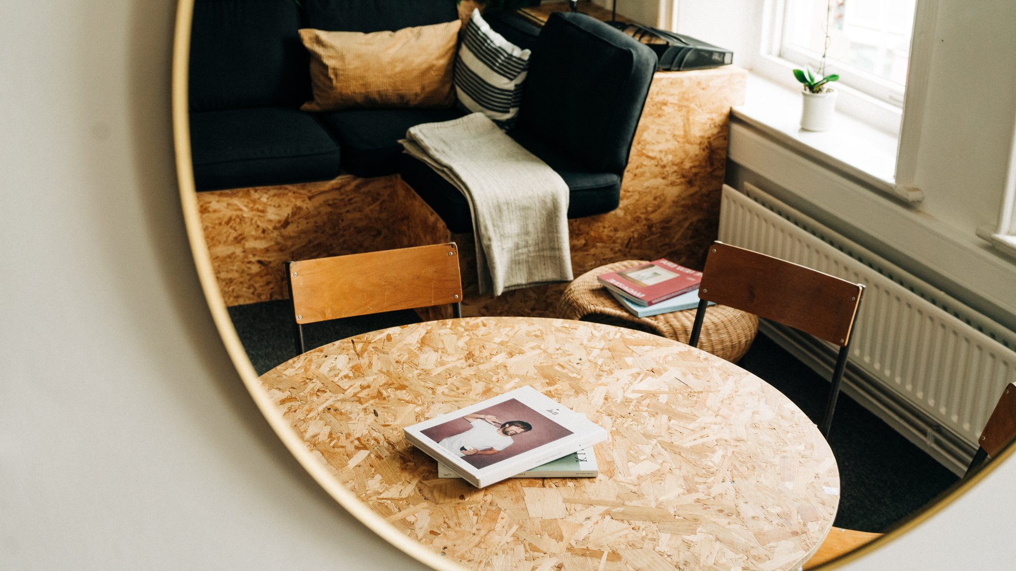 A cozy reading nook with a black sofa, wooden chairs, and a round chipboard table featuring a book on website design with a person on the cover, all reflected in a large circular mirror.