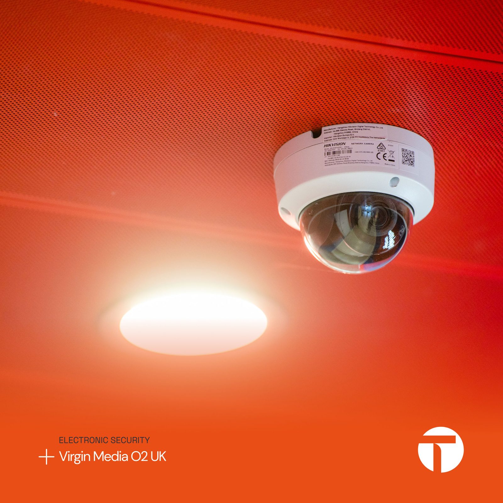 A security camera mounted on a red ceiling next to a bright circular light, with the Virgin Media O2 UK logo indicating electronic security services provided by Brand Agency Cardiff.