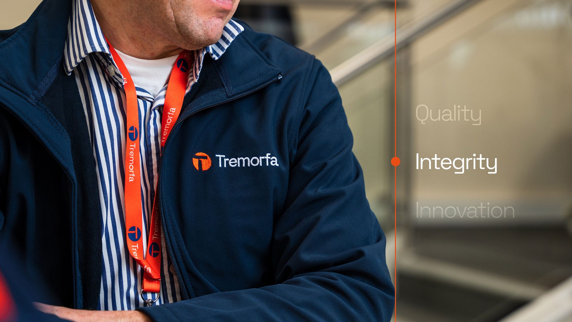 A close-up of a man wearing a blue jacket and striped shirt with a tremorfa lanyard, standing by an escalator. The image includes text on the right reading 