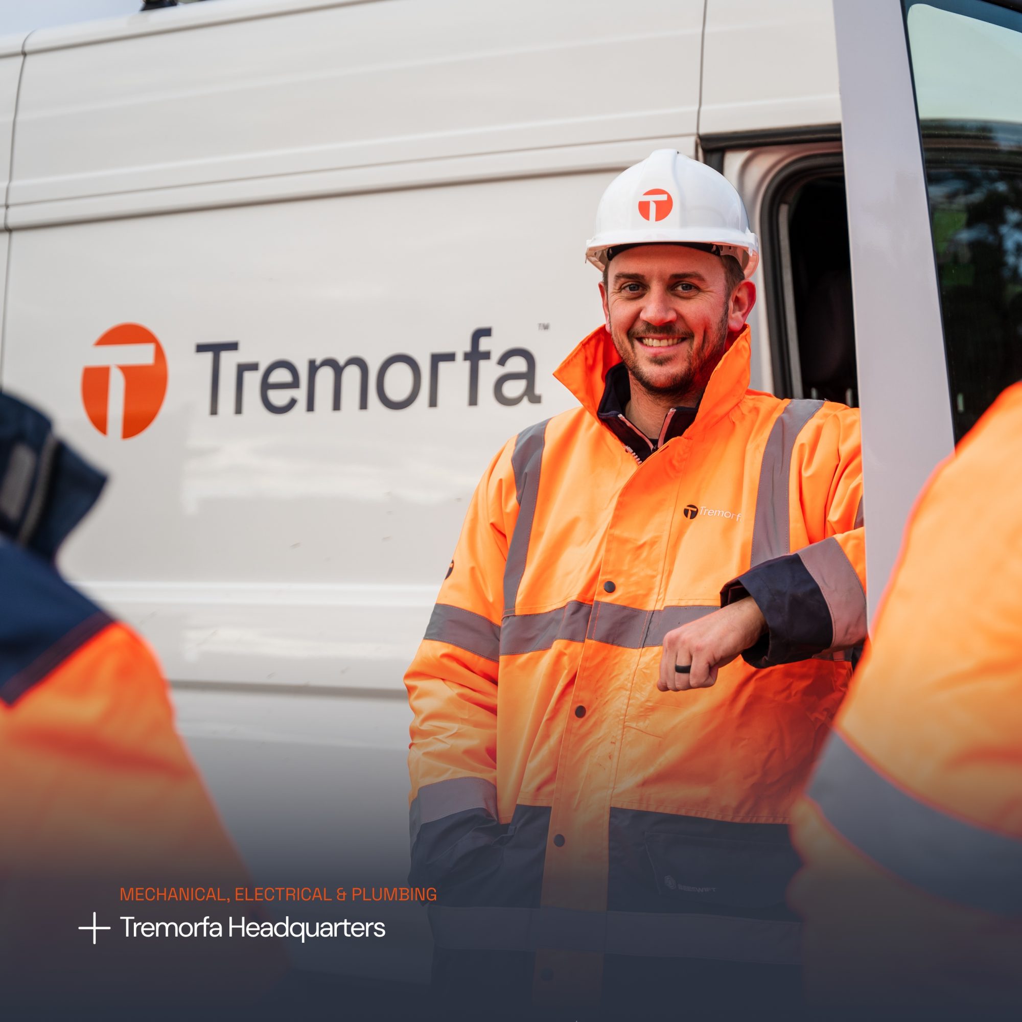 A smiling worker in a high visibility orange jacket and white hard hat stands in front of a white van labeled 