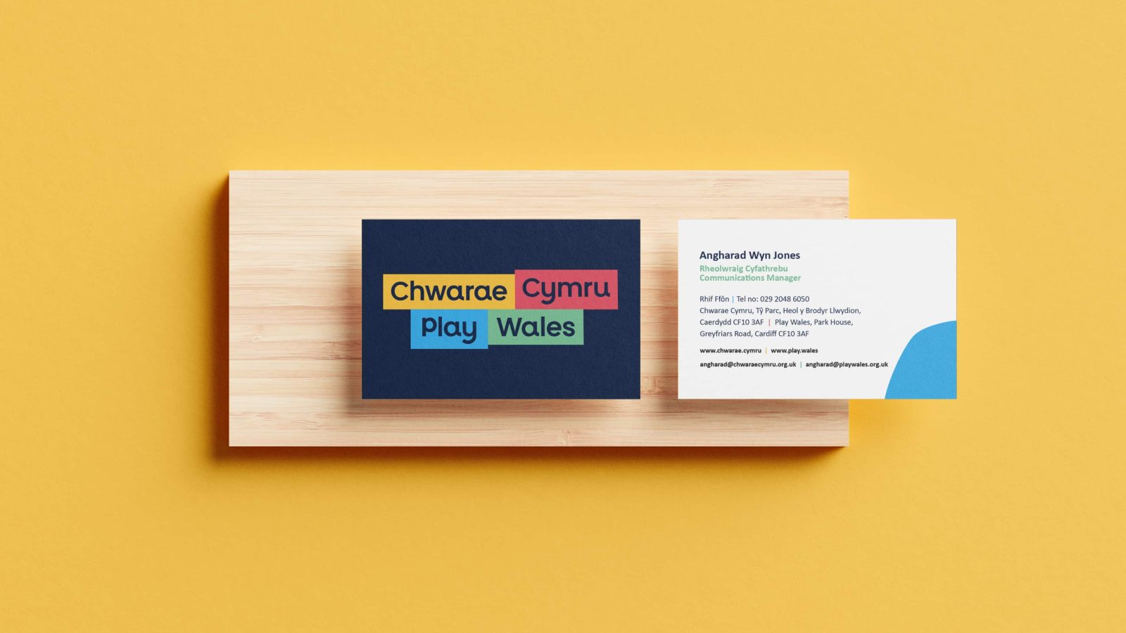 A business card designed by a Brand Agency in Cardiff displayed on a wooden holder against a yellow background. The card features a dark blue design with the logo 