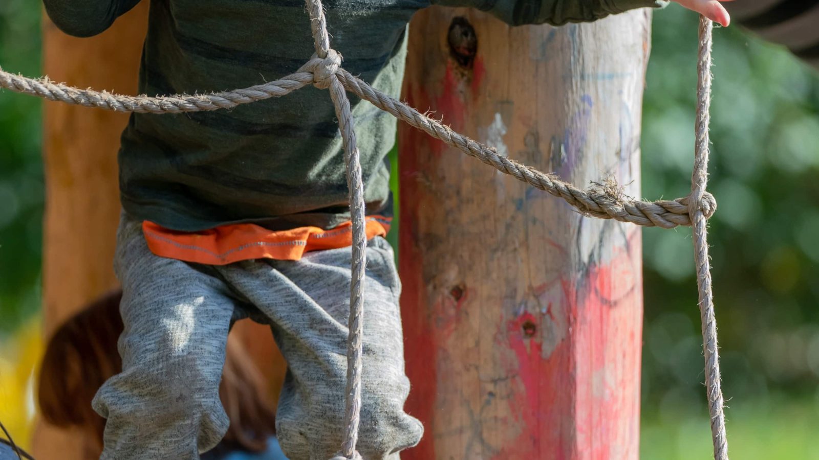 Close-up of a child in gray pants and a green shirt climbing a wooden structure with ropes at an outdoor playground, focusing on the detail and texture of the ropes and wood for a brand strategy portfolio.