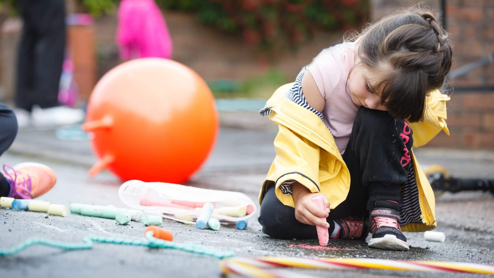A young girl in a yellow raincoat kneels on a driveway, drawing with pink chalk. There are scattered toys and colorful sidewalk chalk around her.