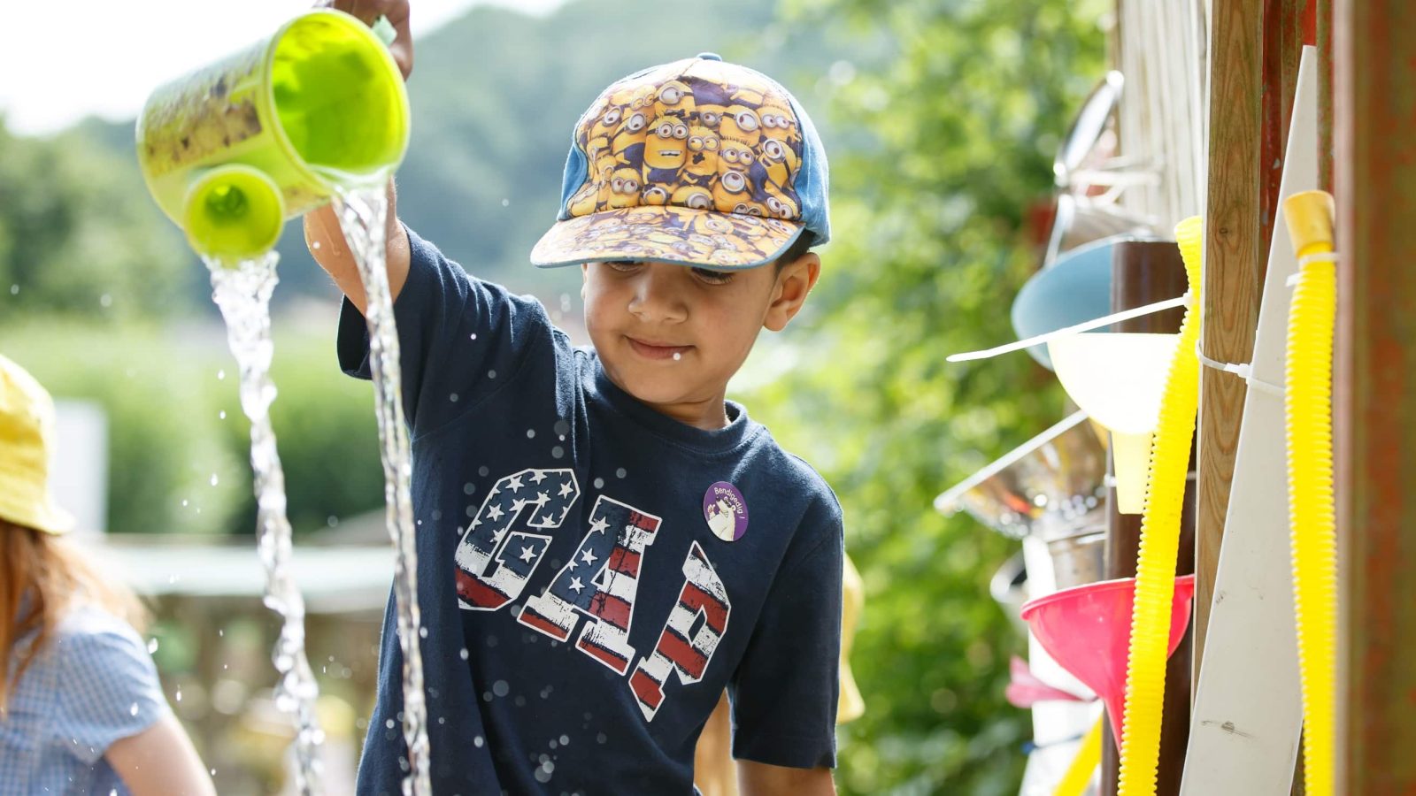 A young boy wearing a minions cap pours water from a green watering can, smiling as he plays outdoors on a sunny day.