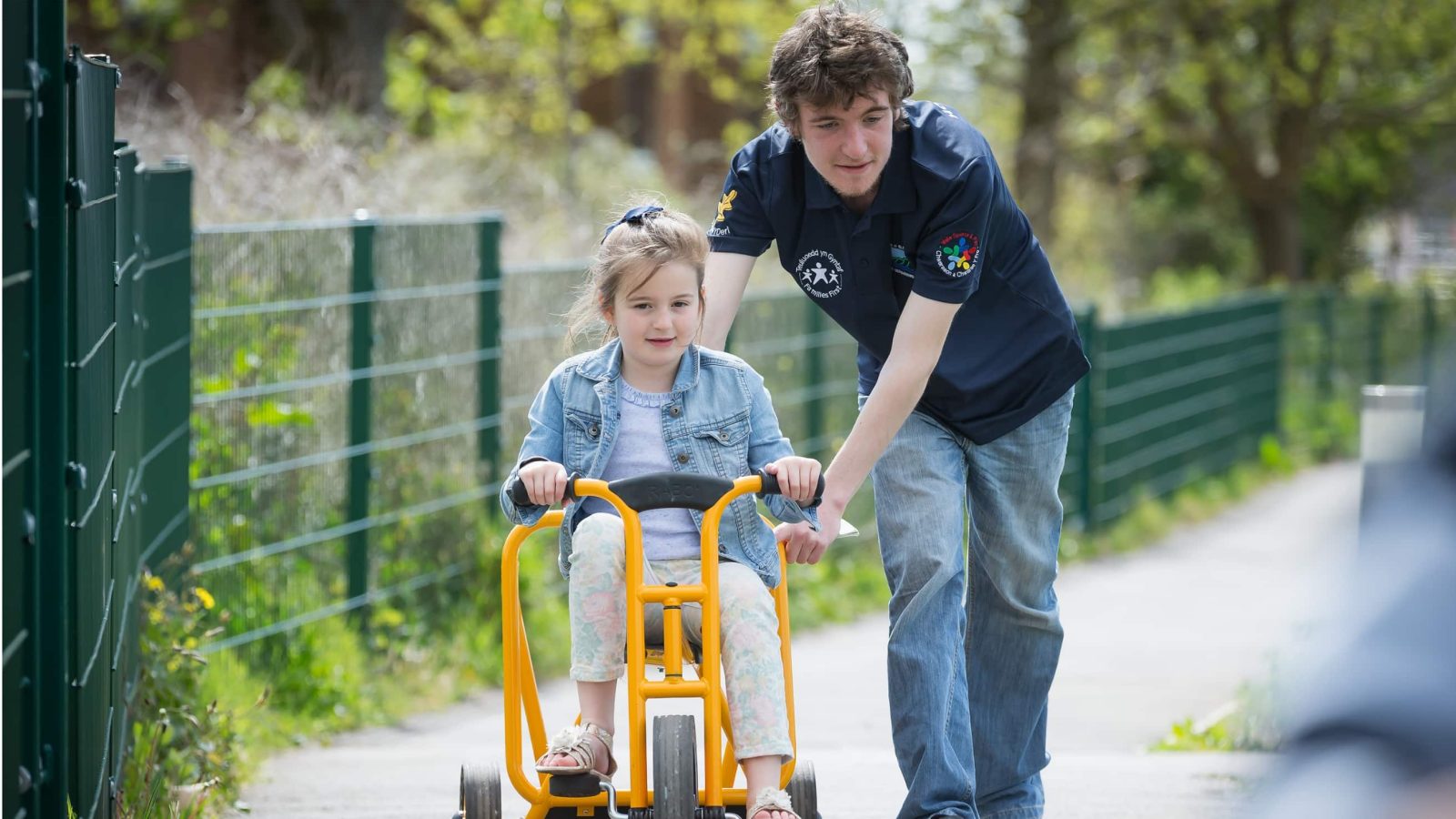 A young girl with a disability using a walker is guided by a male caregiver on a sunny path. She looks focused, the caregiver attentive and supportive, embodying the compassionate image promoted by the Design Agency