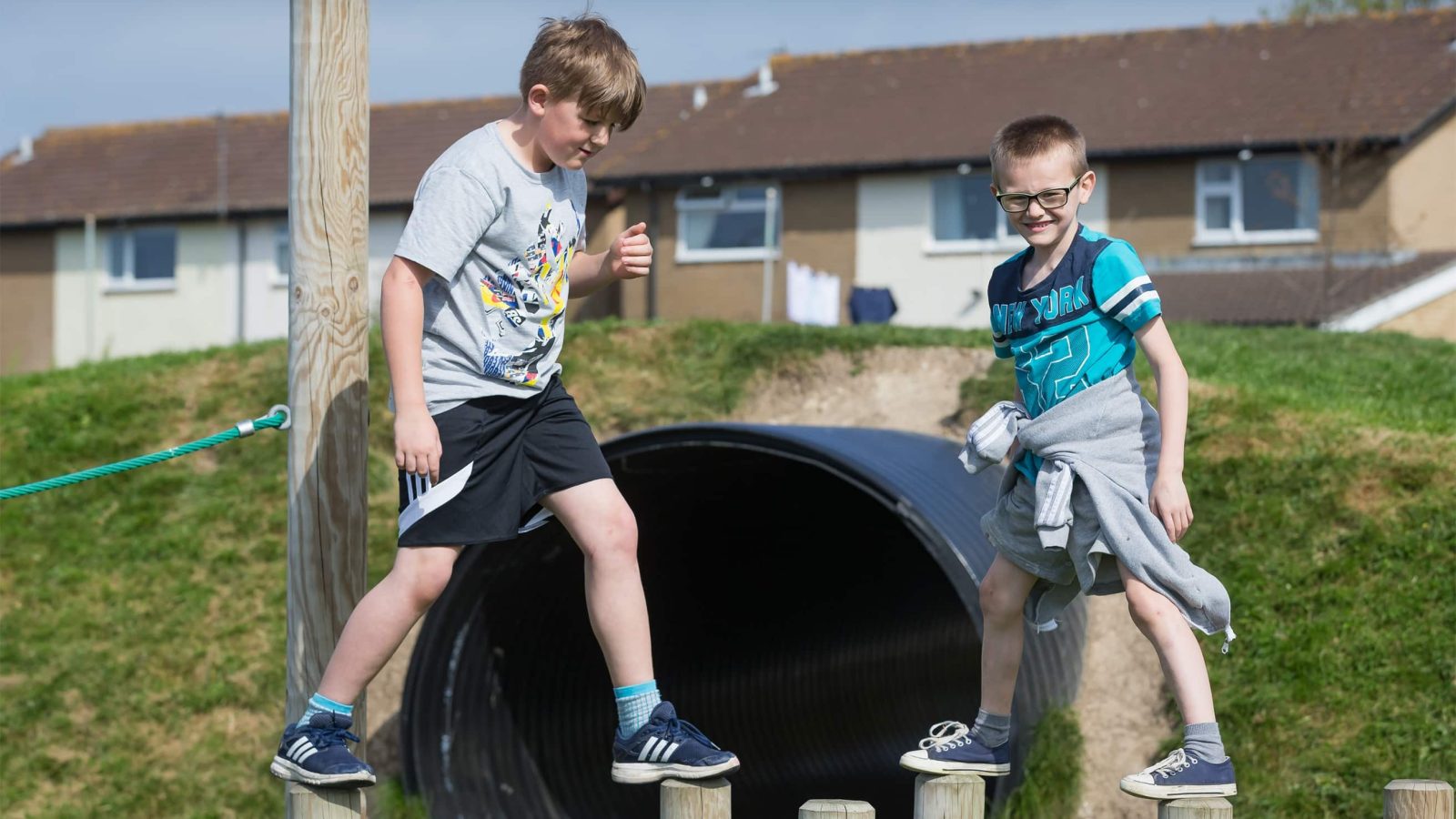 Two boys enjoying a sunny day outdoors on a playground, one standing on a tunnel and the other next to it, both smiling and reflecting the joyful brand strategy of youthfulness.