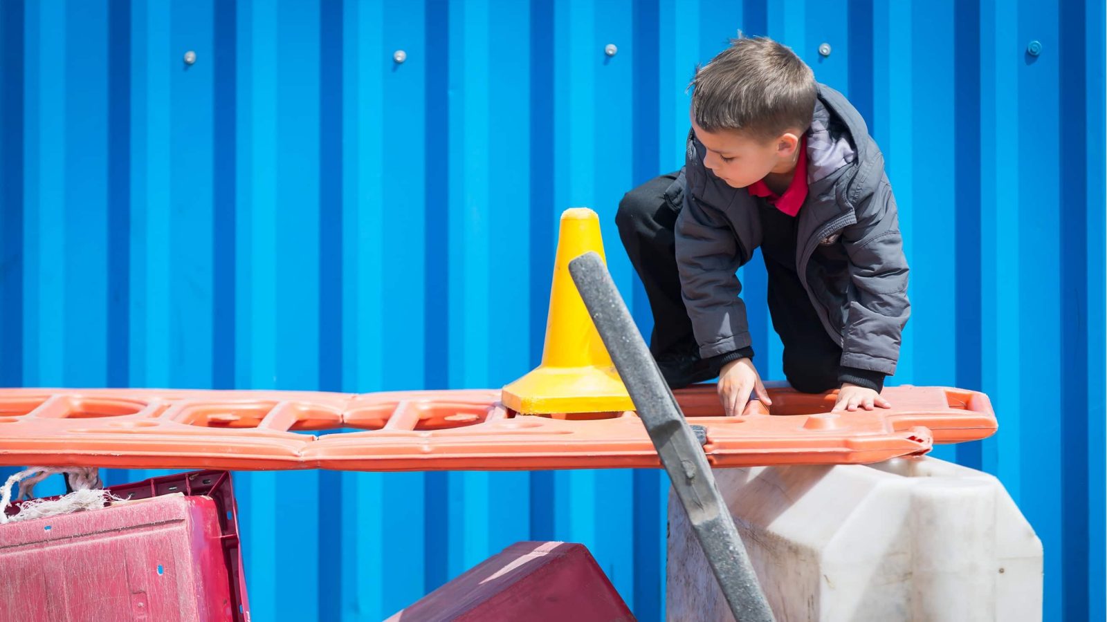 A young boy interacts with a construction barrier in front of a bright blue corrugated metal surface, with a yellow traffic cone visible on the barrier.