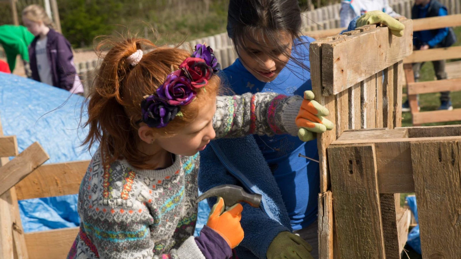 Two young girls, one with a floral headband, engage in building a website design with wooden materials outdoors, using tools and wearing work gloves, with other children in the background.
