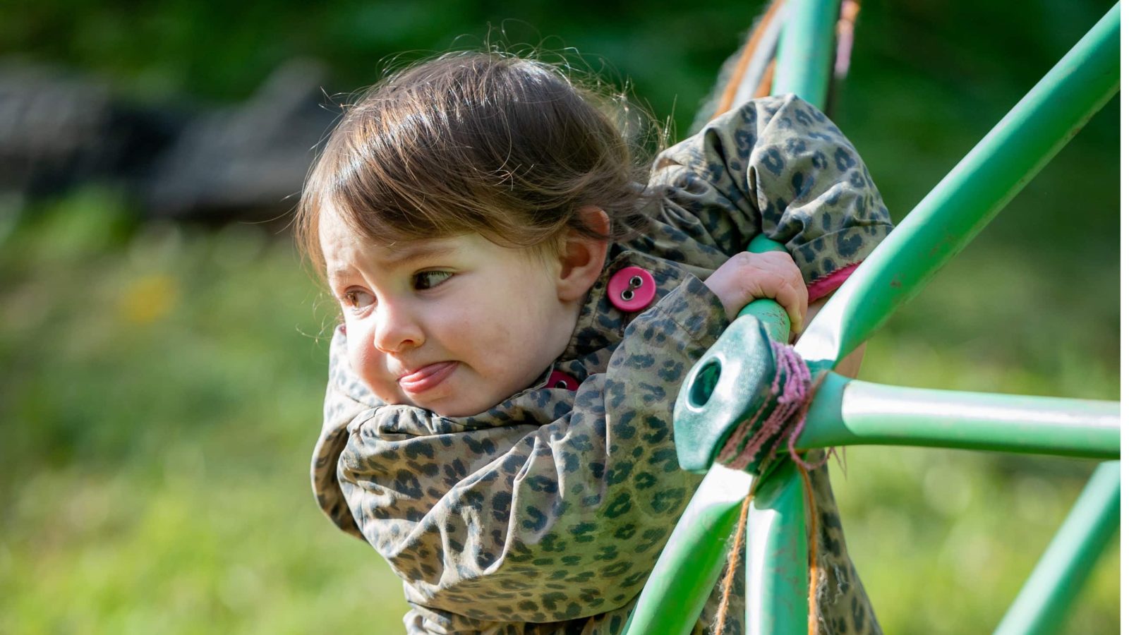 Young girl in a camouflage jacket hugging the bars of a green playground structure, smiling gently with a natural background.
