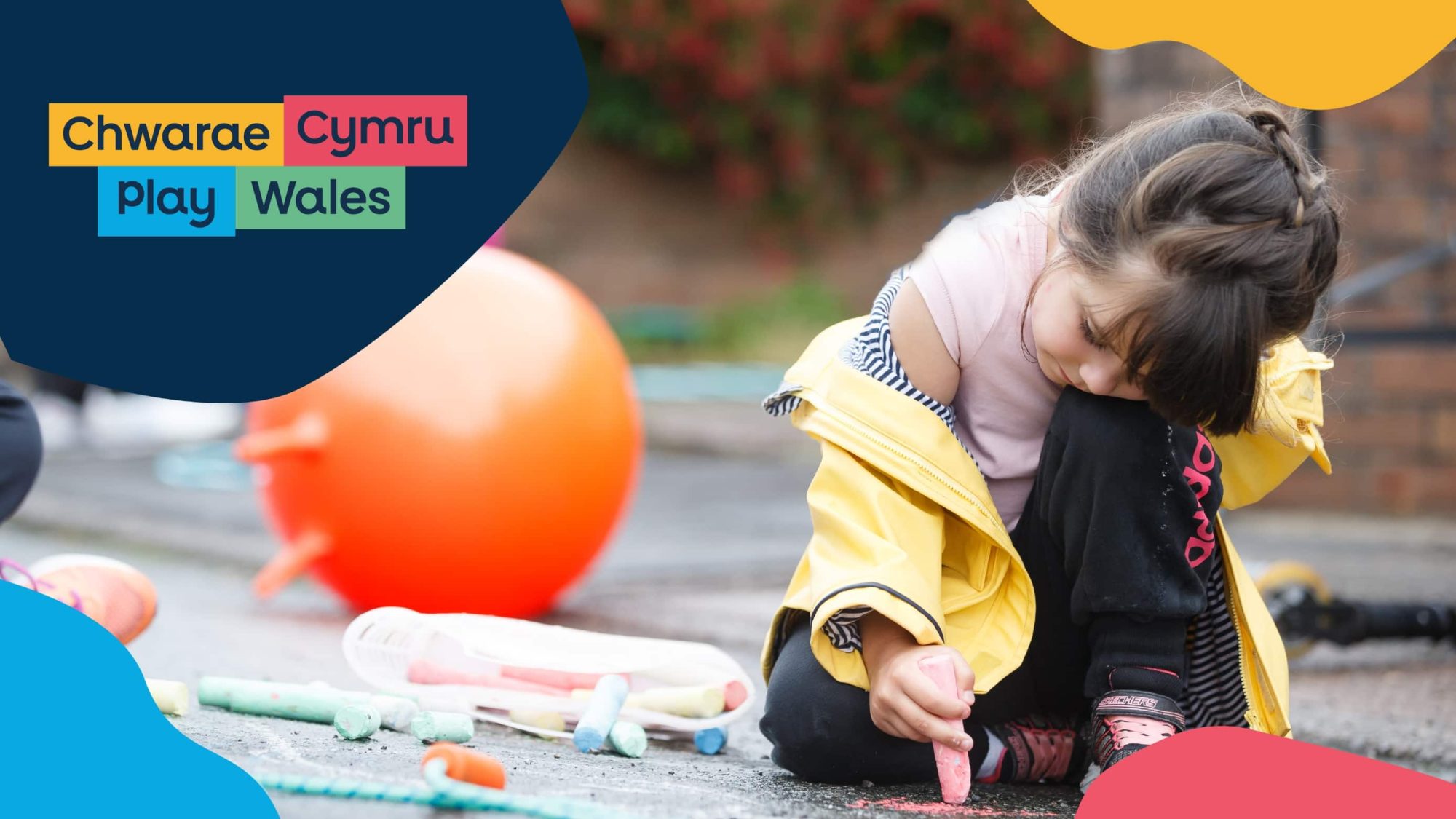 A young girl in a yellow jacket draws with chalk on pavement, surrounded by colorful balls and toys, with the 