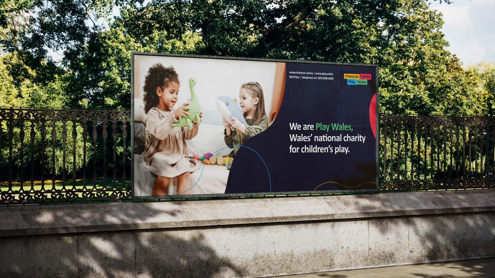 A billboard featuring two children playing happily. the left child, of african descent, holds a green toy, while the right child, caucasian, assists. text promotes play wales, a children’s charity.