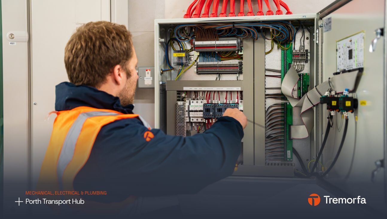 A male technician in an orange vest is working on an electrical panel with numerous wires and components, in a labeled environment suggesting it's related to mechanical, electrical, and plumbing services at a design agency.