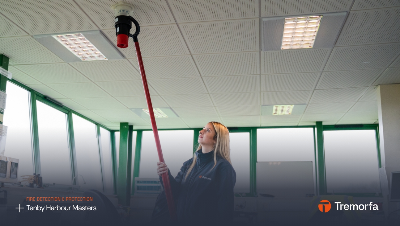 A woman in a dark uniform uses a long red tool to test a ceiling-mounted fire detector in a bright office space with the logo of Tenby Harbour Masters and Tremorfa on it, updated by