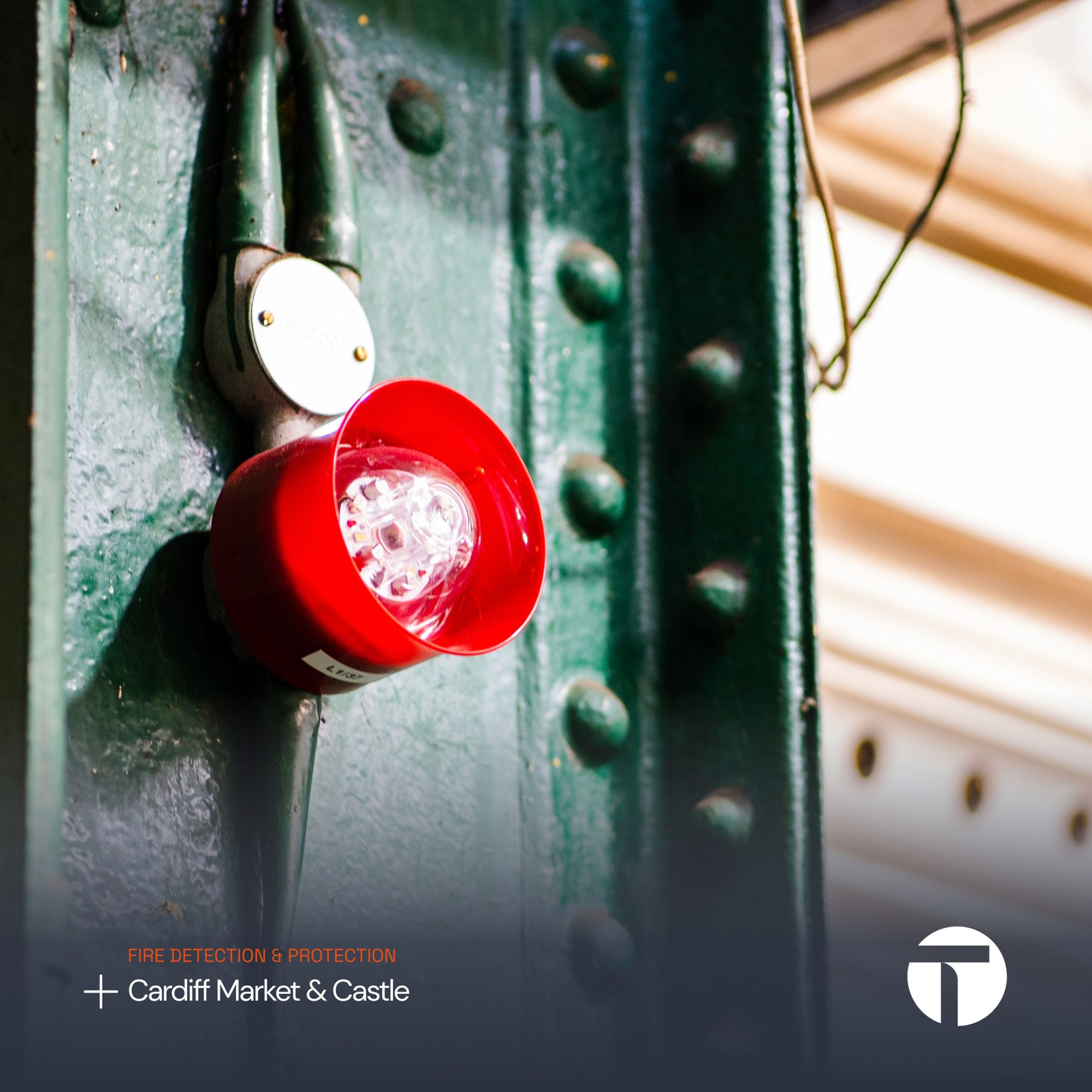 A close-up of a red fire alarm light mounted on a green textured wall, with subtle text referencing fire protection services in Tremorfa market & castle.