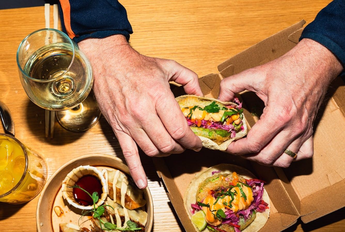 A person holding a taco over a wooden table, where alongside sits a mysterious 