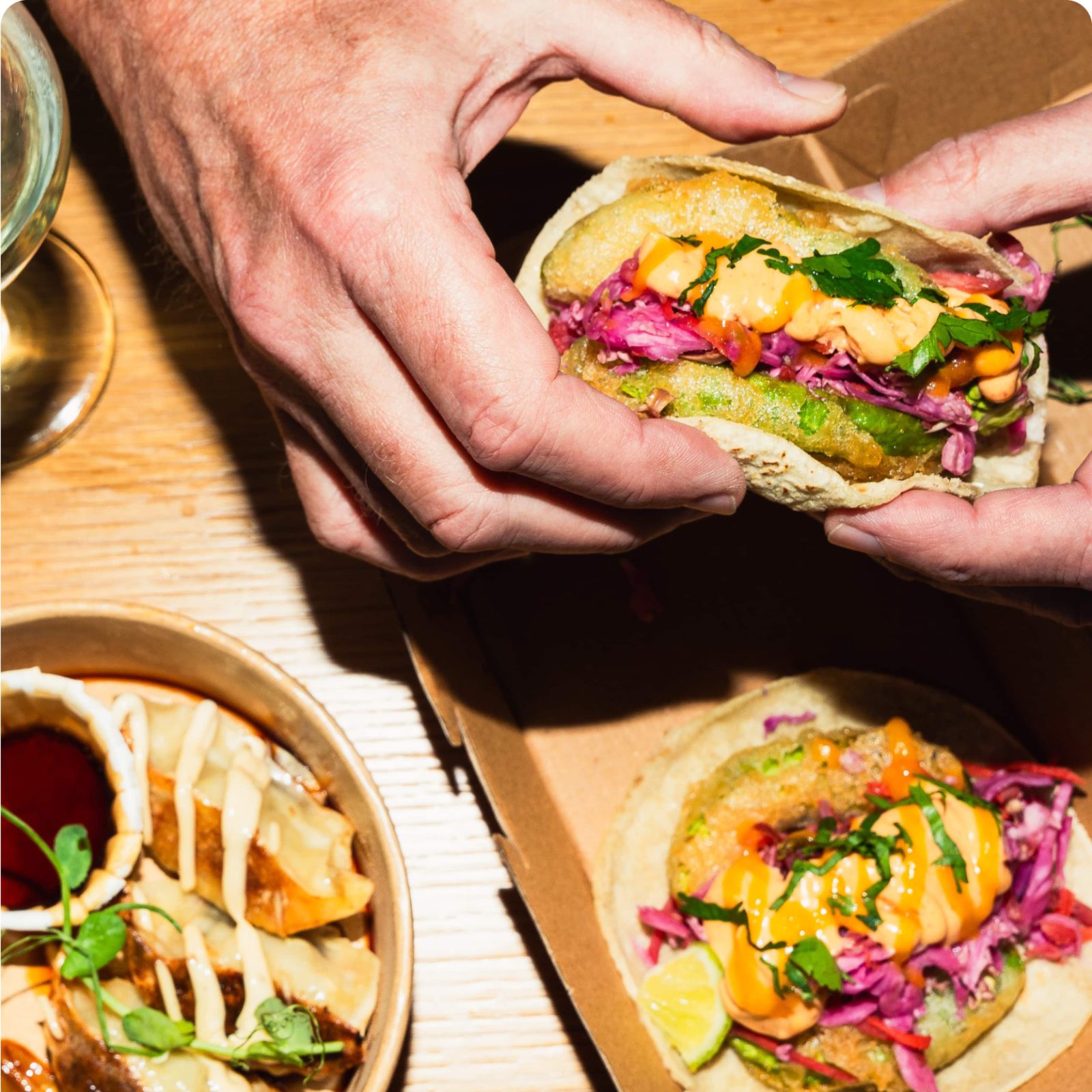 Hands hold a delicious taco brimming with toppings, sitting next to a dish of dumplings and sauce on a wooden table. The scene feels like a fusion feast that even the big, bad wolf wouldn't resist.