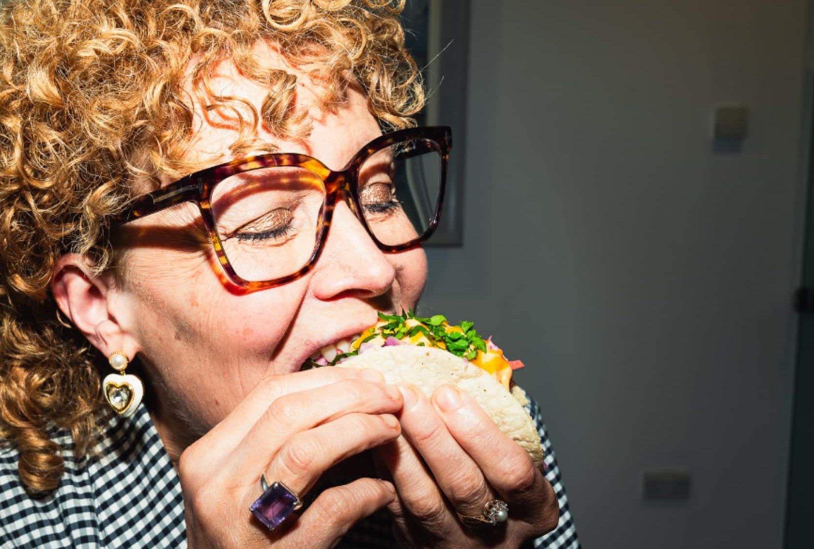 In a dimly lit environment, a person wearing glasses and checkered clothing bites into a sandwich with both hands, embracing their inner bad wolf.