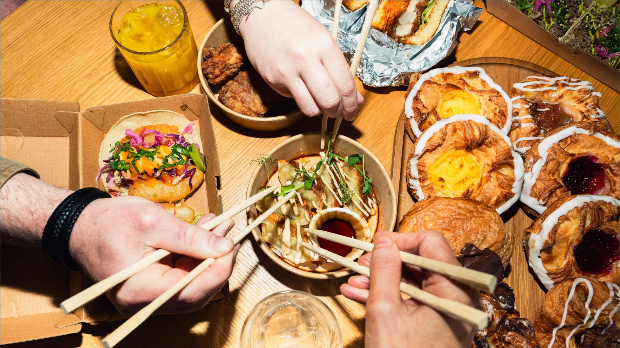 Several people use chopsticks to pick food from bowls on a table, surrounded by pastries and an array of drinks. The scene feels like a gathering before the bad wolf, savoring moments of delight and camaraderie.