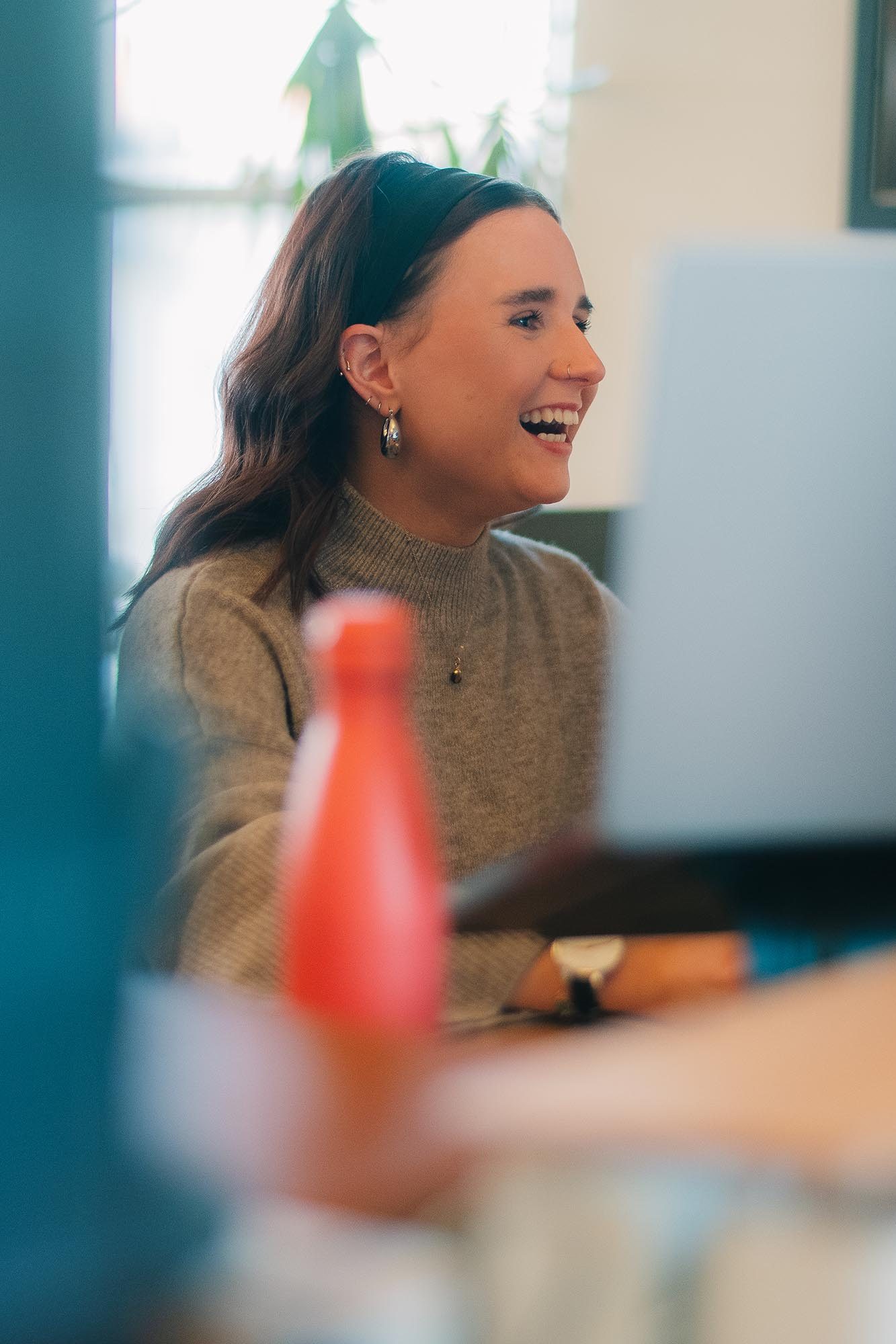 A woman in a gray sweater smiles while working at a computer in an office setting at Brand Labs. A red bottle is on her desk.