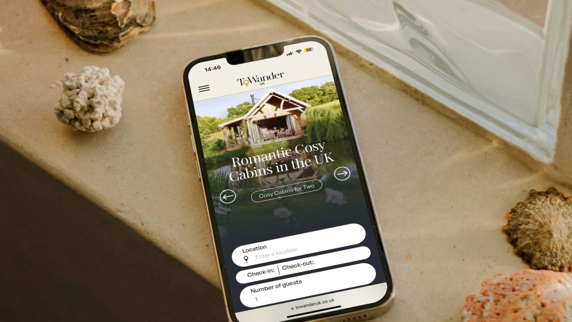 A smartphone on a windowsill, surrounded by seashells and rocks, displays the homepage for Bad Wolf romantic cabins in the UK.