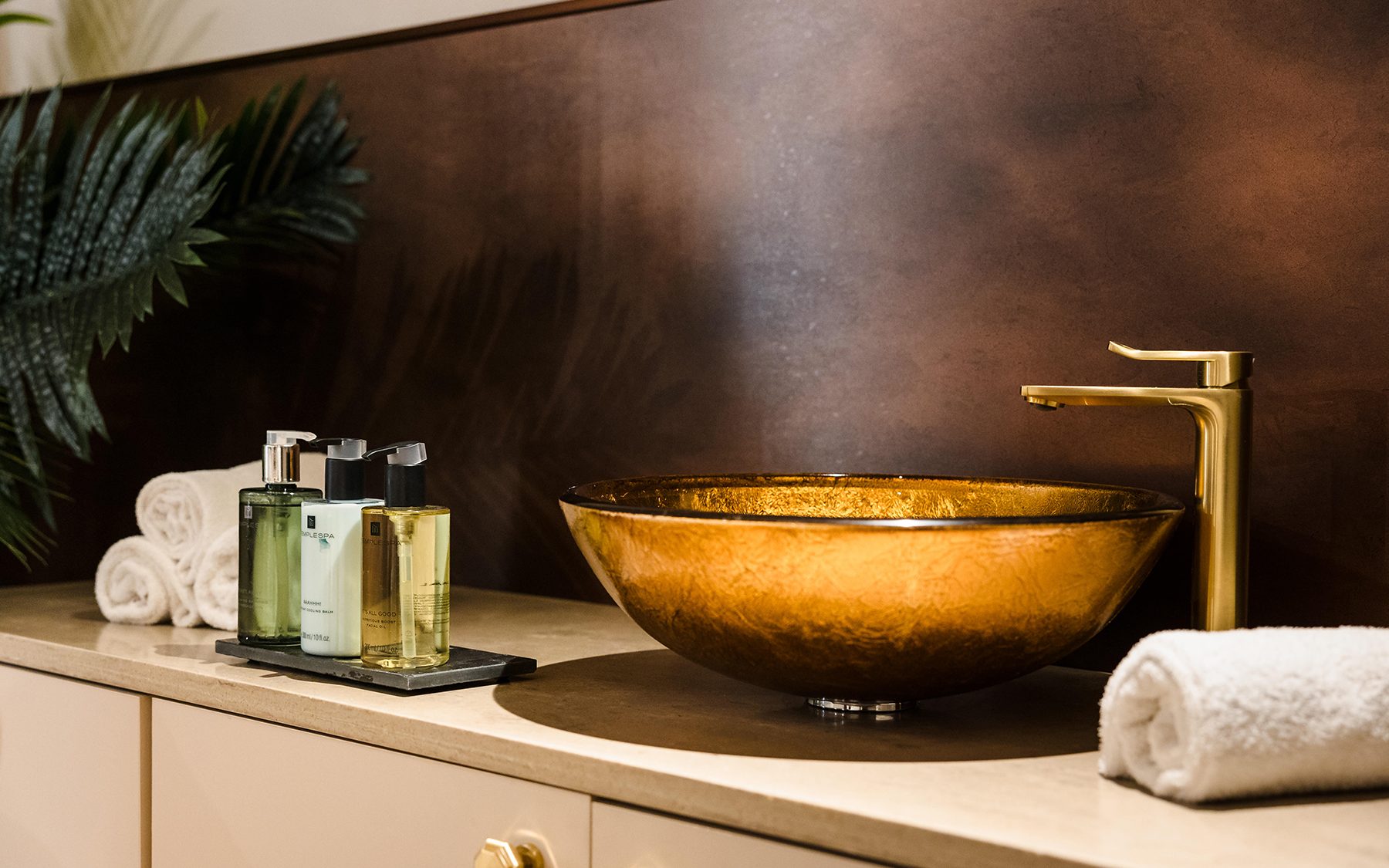 A bathroom sink with a gold basin and faucet, toiletries, and neatly rolled white towels on a beige countertop.