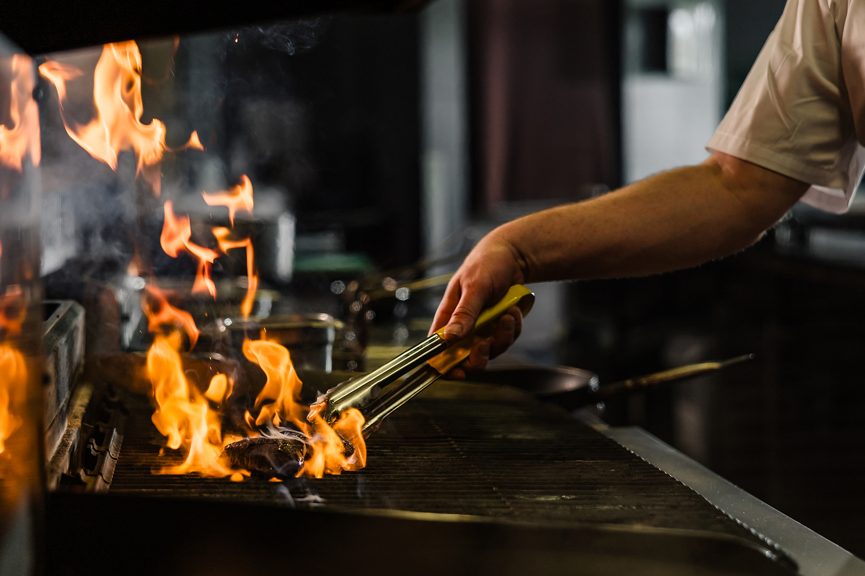 Chef grilling meat on a flaming grill using tongs, with pots in the background.