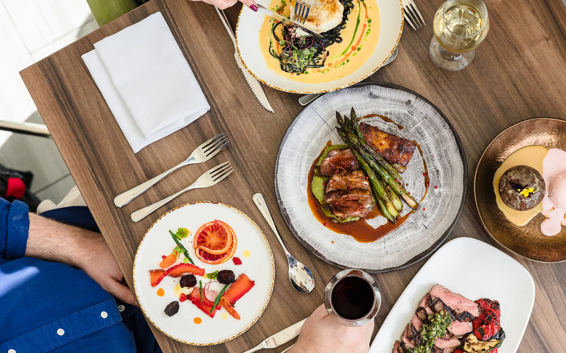 Aerial view of a dining table with plated gourmet meals, including meat dishes, salad, dessert, and drinks.